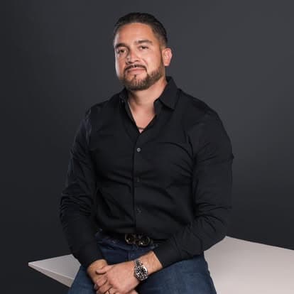 Efrain Gomez Jr. in a dark shirt, seated and looking at the camera in a studio portrait