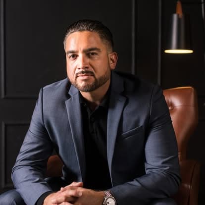 Efrain Gomez Jr. wearing a suit jacket, seated in a leather chair against a dark paneled wall