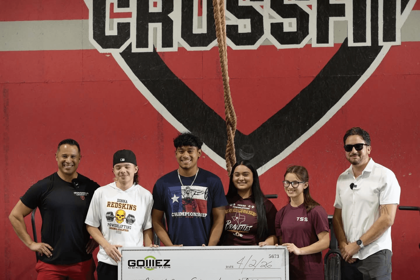Efrain Gomez Jr. and a coach standing with four student athletes holding an oversized Gomez Construction scholarship check at a gym