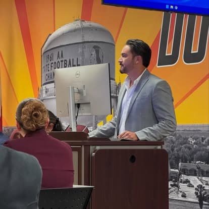 Efrain Gomez Jr. speaking from a lectern at a community meeting, with a Donna water tower mural on the wall behind him